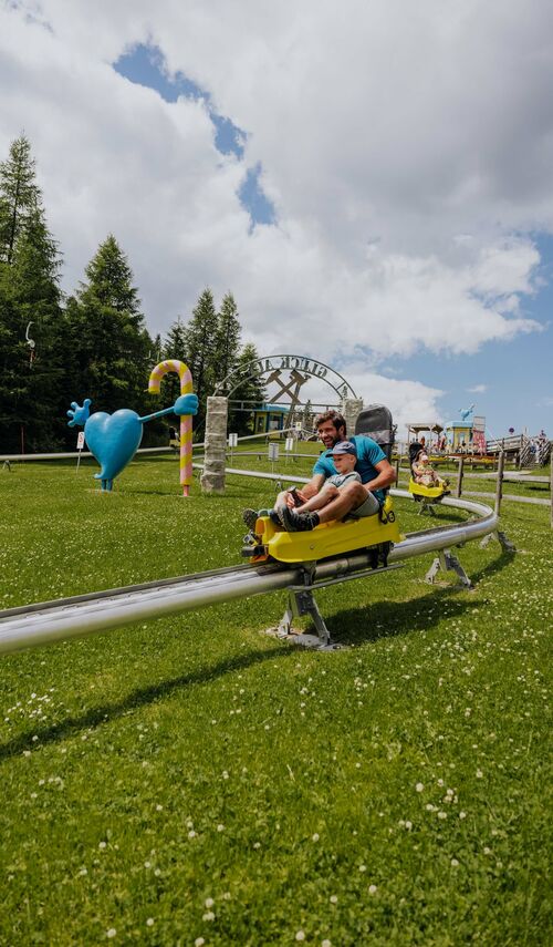 das Bild zeigt einen Vater mit seinem Sohn auf der Sommerrodelbahn, der Himmel ist etwas bewölkt