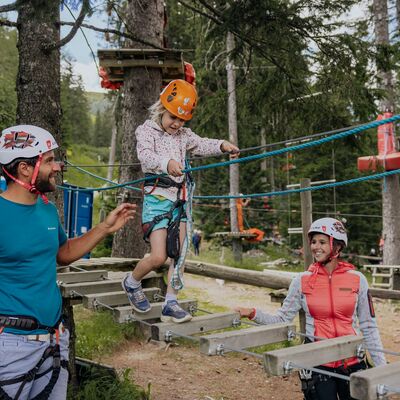Eine Familie klettert im Hochseilgarten, gut gesichert mit Helmen und Gurten