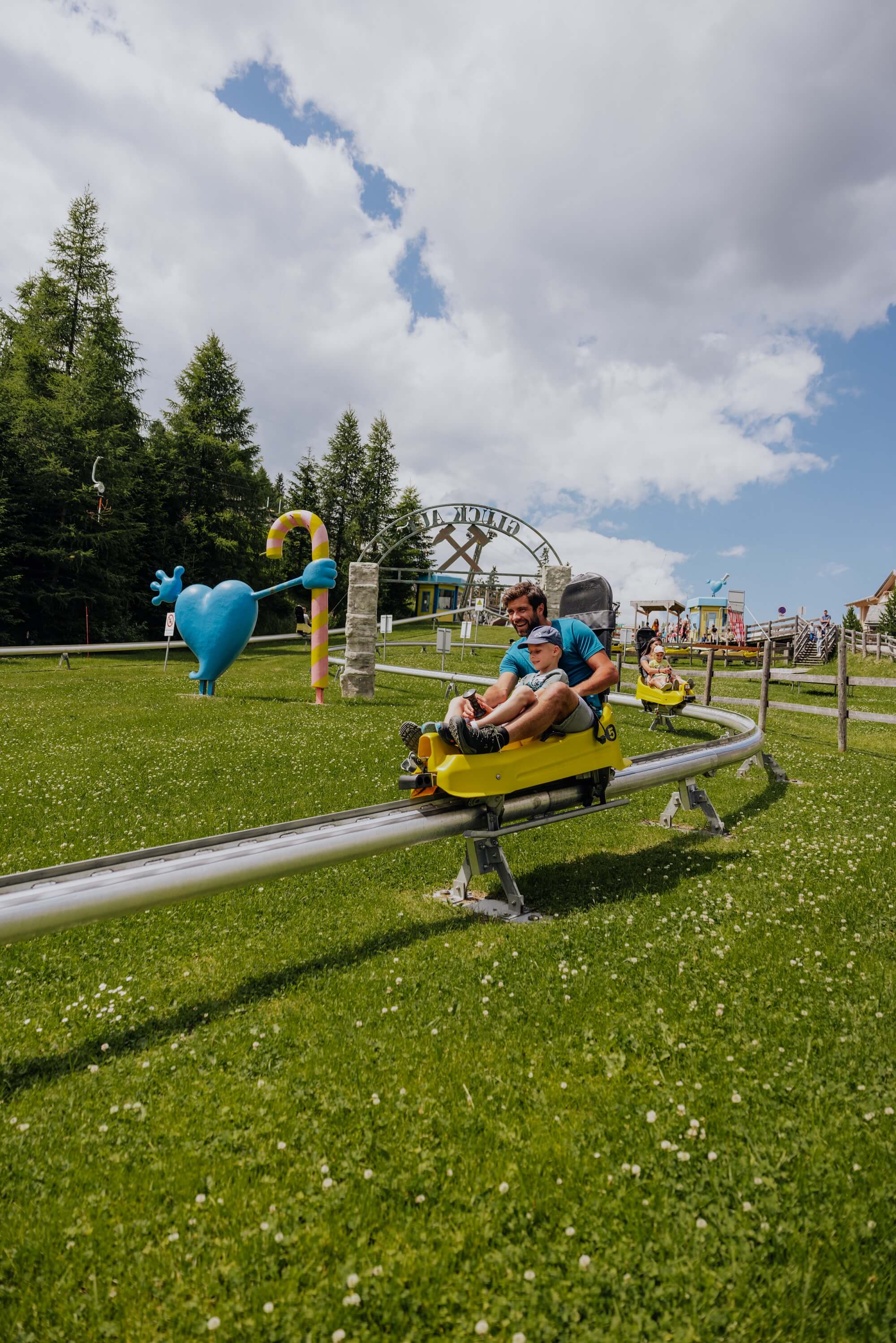 das Bild zeigt einen Vater mit seinem Sohn auf der Sommerrodelbahn, der Himmel ist etwas bewölkt